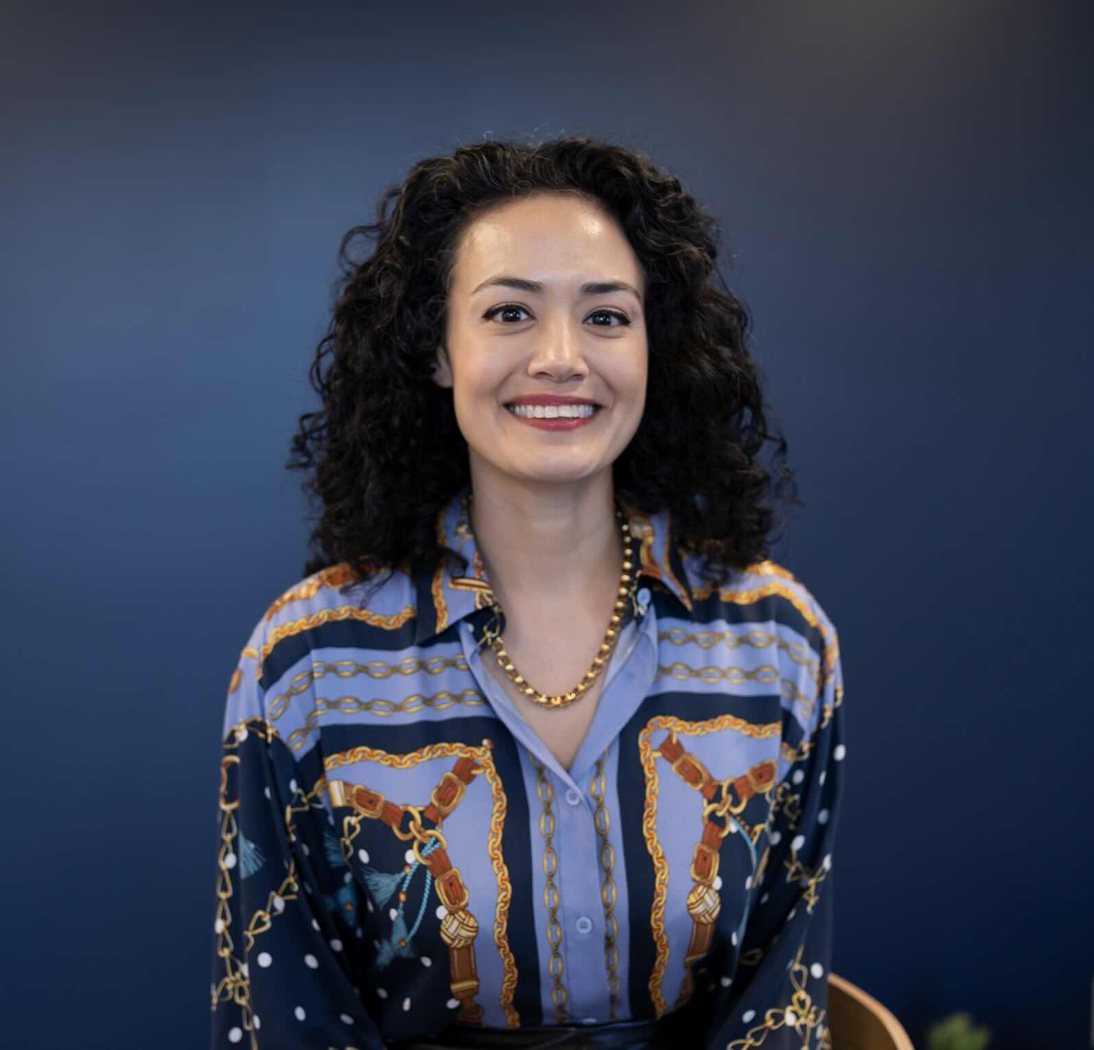Whitney Masulis, smiling, wearing a patterned blue blouse against a dark blue background